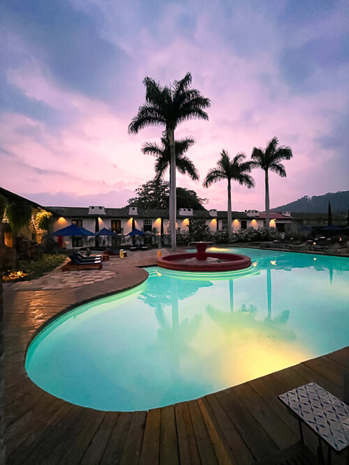 a large illuminated pool with tall palm trees and pink sunset sky as a backdrop