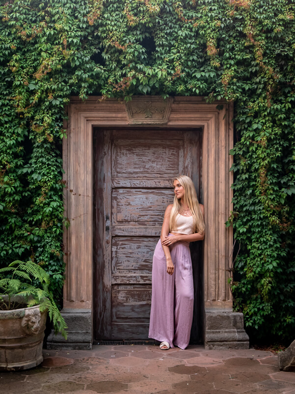A woman leaning on an old wooden doorway surrounded by greenery