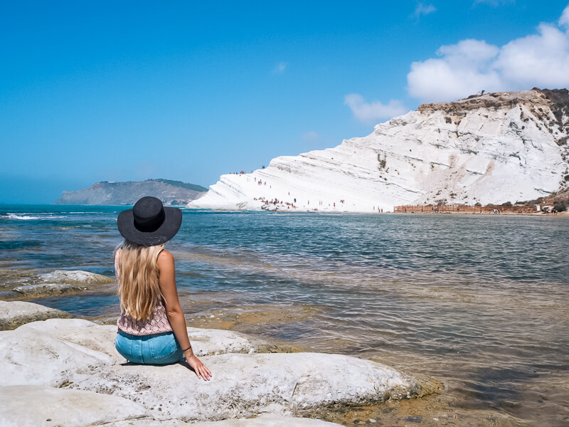 A woman sitting on a rock looking at the white marl cliff of Scala dei Turchi on Sicily's southern coast, a must-visit on every Sicily road trip.