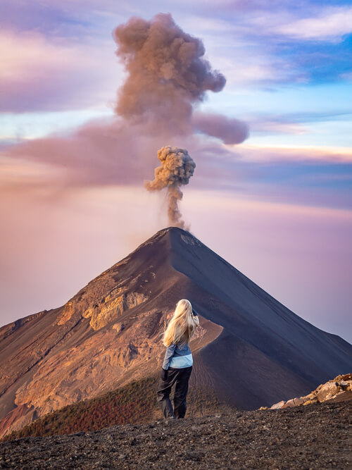 A woman looking at a smoking volcano during sunrise on the Acatenango volcano trek, one of the highlights of Guatemala