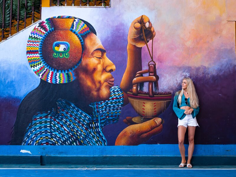 A woman leaning on a wall displaying a colorful mural of a Mayan man in San Juan, Lake Atitlan