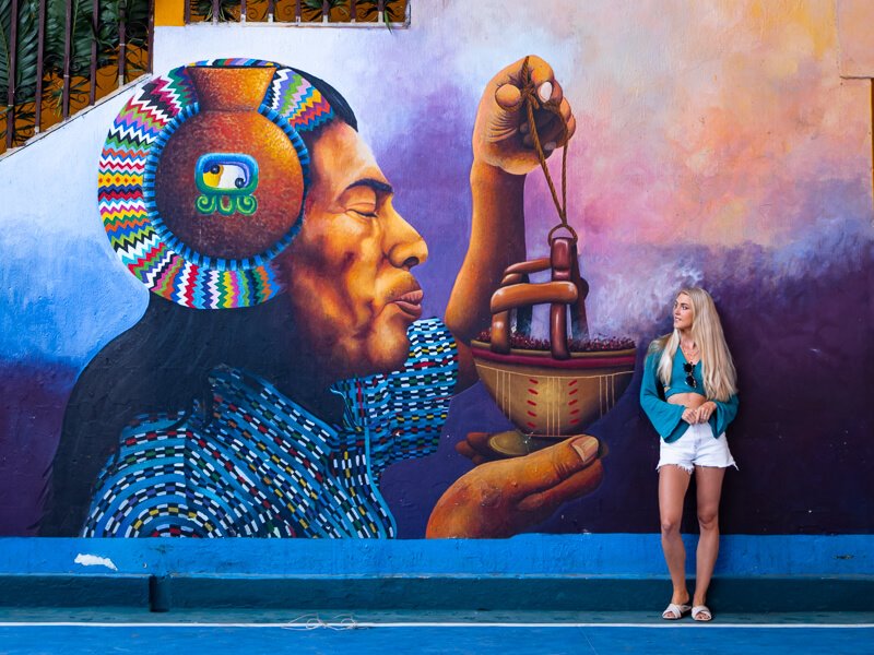 Me leaning on a wall displaying a colorful mural of a Mayan man in San Juan, Lake Atitlan