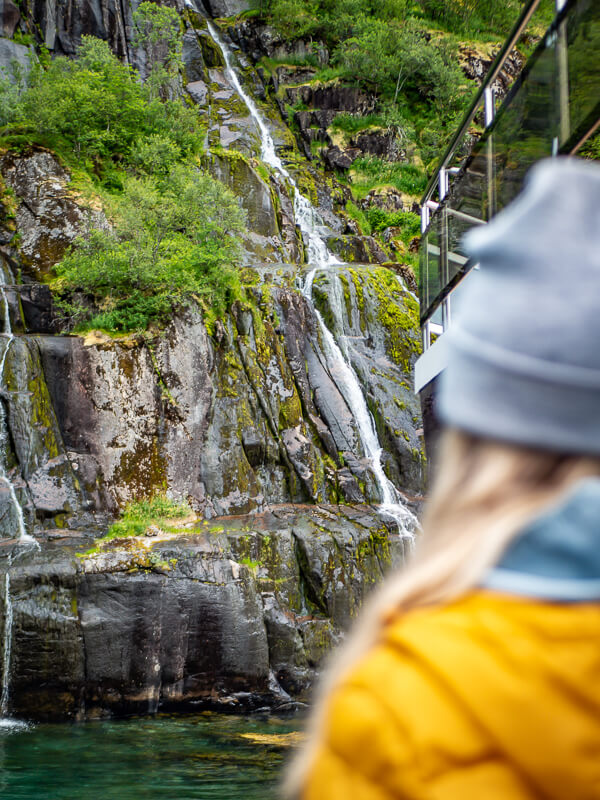 A woman standing on a ship looking at a small waterfall surrounded by green vegetation on a Trollfjord cruise in Lofoten