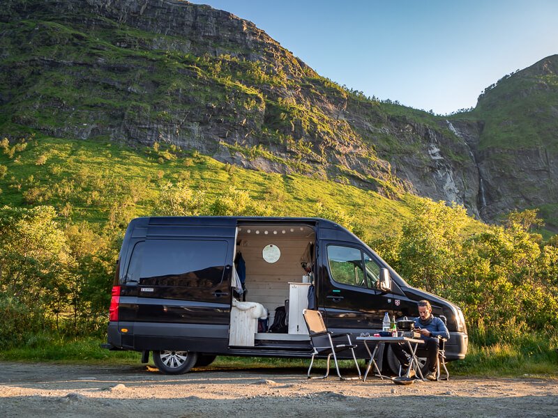 A man sitting on a camping chair in front of a black campervan in Norway