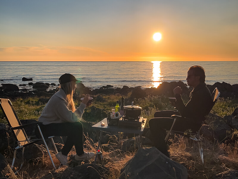 A man and a woman having a camping dinner on a rocky beach with sunset view on the Lofoten Islands