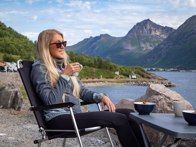 A woman sitting on a chair next to a scenic fjord while wild camping in Norway with a campervan