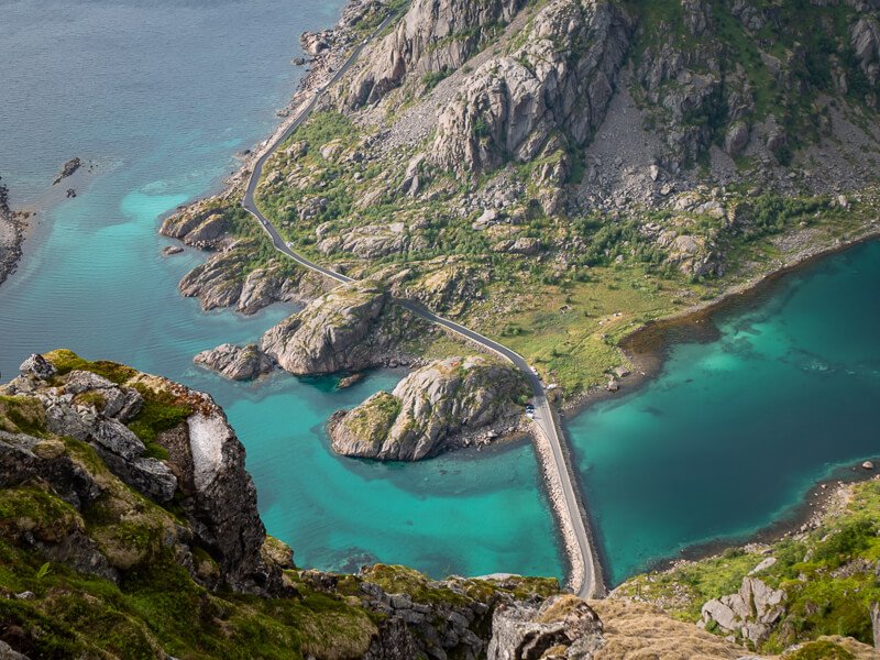 A narrow coastal road in Norway, surrounded by mountains, rocky islets and bright turquoise sea