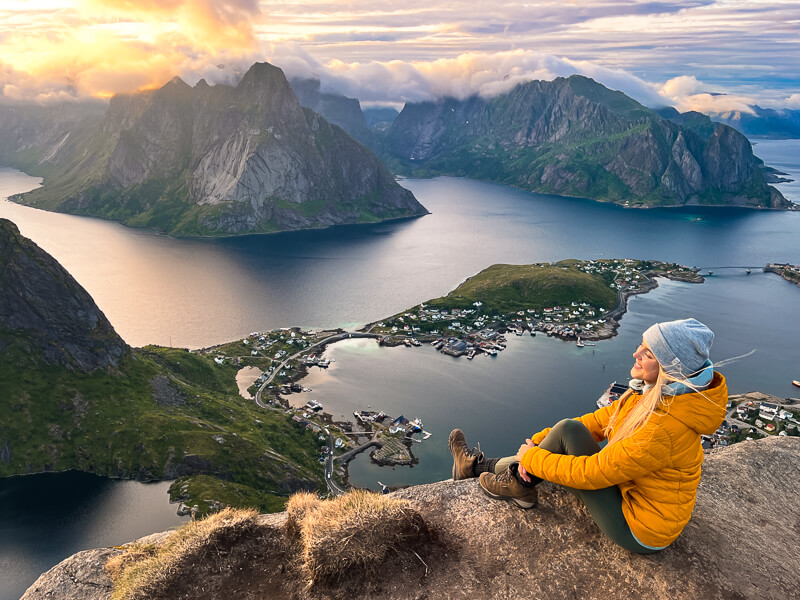 A woman sitting on a rock overlooking a picturesque fjord at Reinebringen peak, one of the best hikes in Lofoten Islands