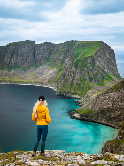 A woman in a yellow jacket standing on the edge of a cliff, overlooking the dramatic mountains of Værøy island