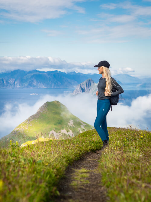 A woman standing on a trail on a meadow with a backdrop of jagged mountains and low clouds on Værøy island in Lofoten