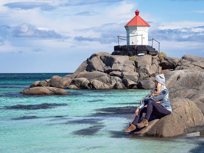 A woman sitting on a rock surrounded by bright blue sea and a small lighthouse in the background at Eggum