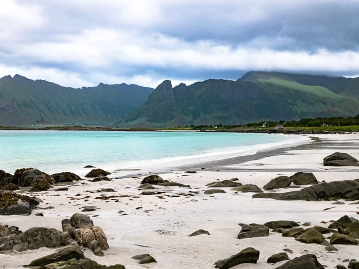 Brown rocks, white sand and a view of  green mountains in the distance at Gimsøy Beach, a hidden gem in Lofoten 
