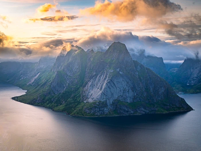 A jagged mountain with sharp peaks rising out of Reinefjord with colorful clouds above it at sunset