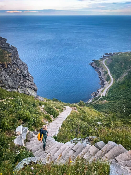 A woman walking up steep sherpa stairs at the Reinebringen hike with a view of the sea in the background