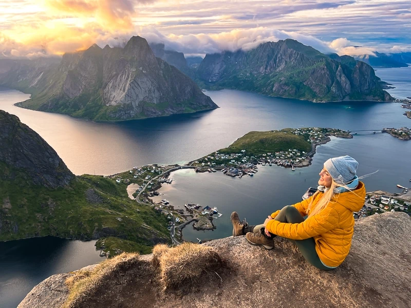 a woman in a yellow jacket sitting on a rock overlooking a fjord surrounded by mountains at the Reinebringen hike, one of the best hikes on the Lofoten Islands