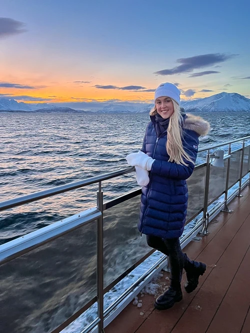 A woman standing on the deck of a whale safari boat with mountains and orange sky in the background