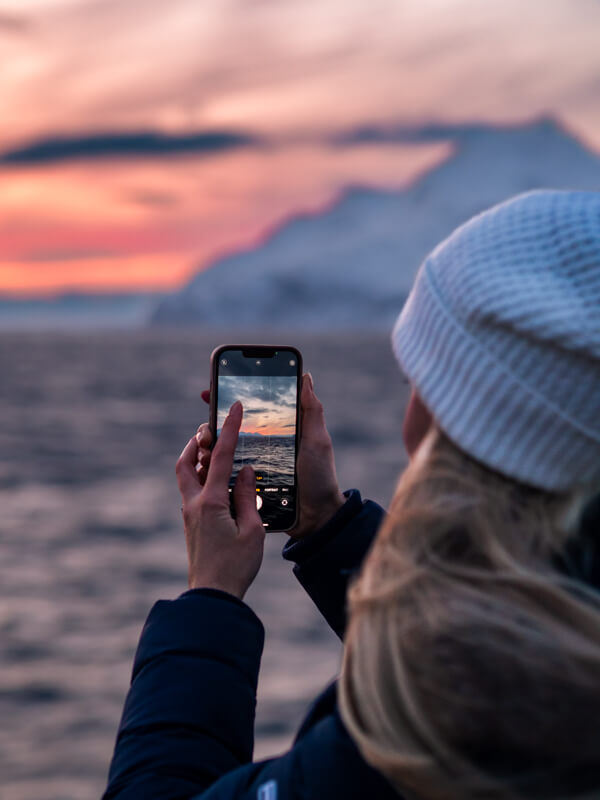 Me holding a phone and taking a photo of the pink twilight sky and snow-covered landscapes on our Tromso whale watching tour.