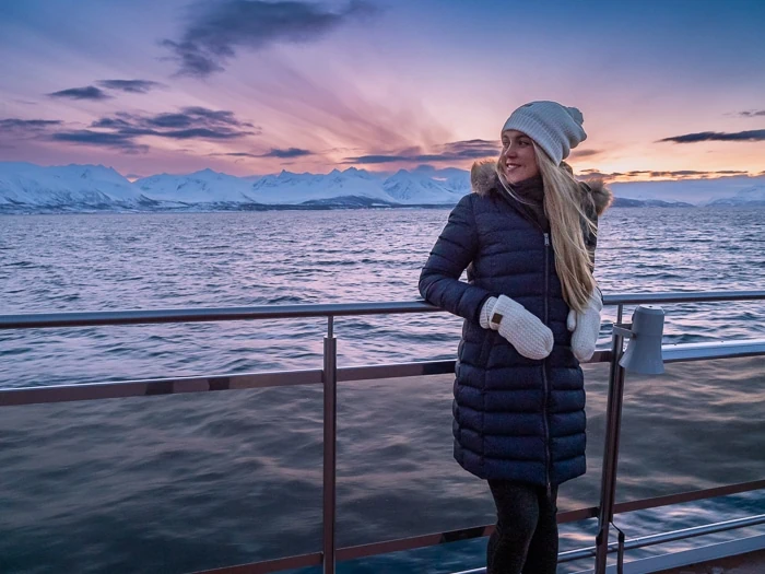 A woman posing on the outdoor deck of a boat with picturesque Arctic landscapes and colorful sky in the background