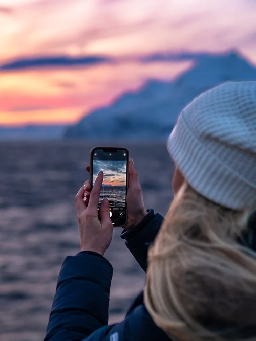 Me holding my phone to take photos of the snowy landscapes and pink skies on our whale watching tour in Tromso.