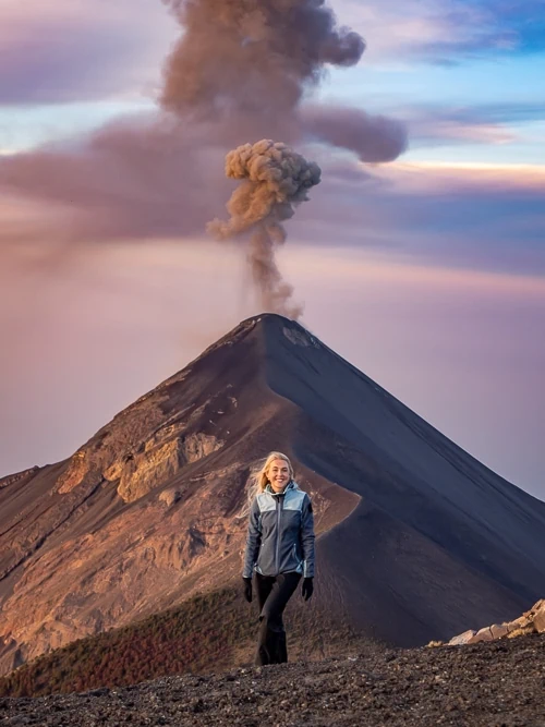 A woman standing on the summit of Acatenango Volcano with the smoking Volcan Fuego in the background