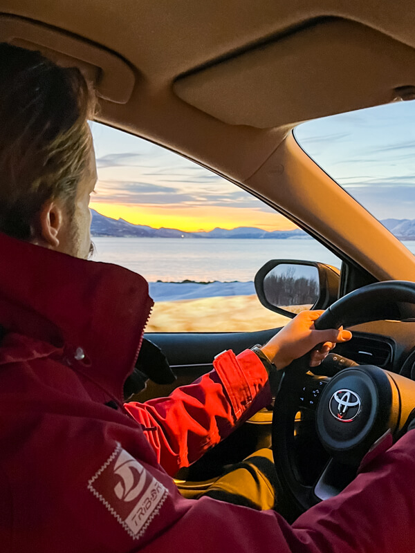 My husband driving our rental car along a fjord in Tromso, with snowy mountains and colorful twilight sky outside the car windows.