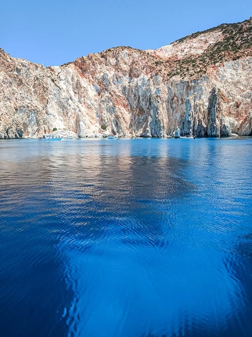 A bay with clear azure water surrounded by cliffs and volcanic rock formations in shades of orange, red and white at Polyaigos Island