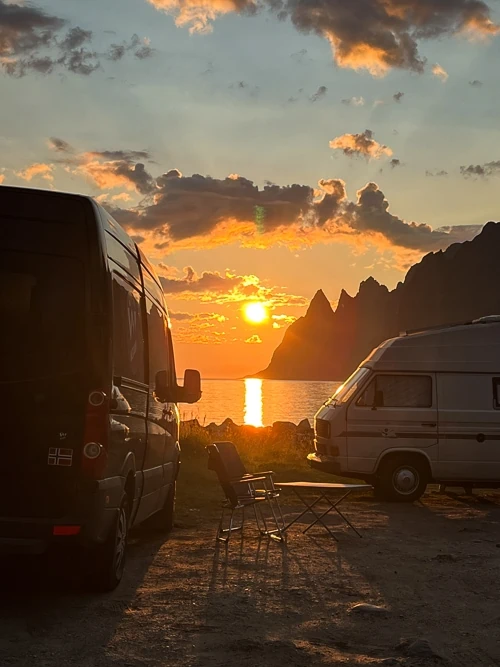 two campervans parked next to a fjord with a view of the Midnight Sun in the background