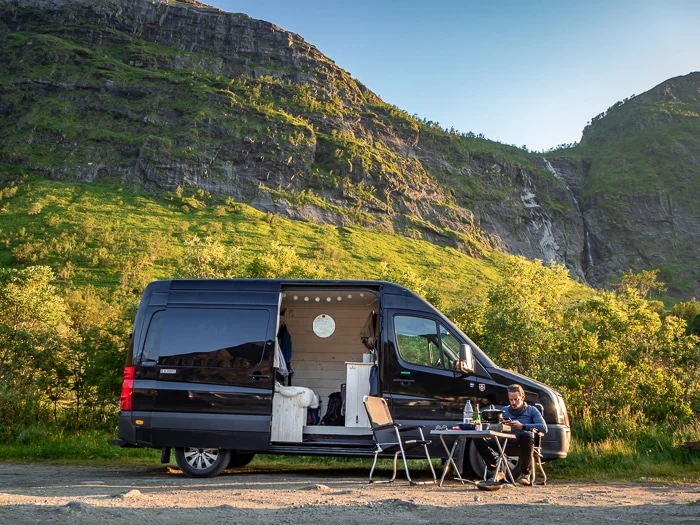 A man sitting on a camping chair in front of a black campervan parked next to a mountain