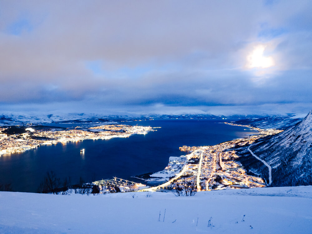 A view of the city lights of Tromso from Storsteinen Mountain, with snow-capped mountains in the background under a cloudy sky with the moon peeking through the clouds.