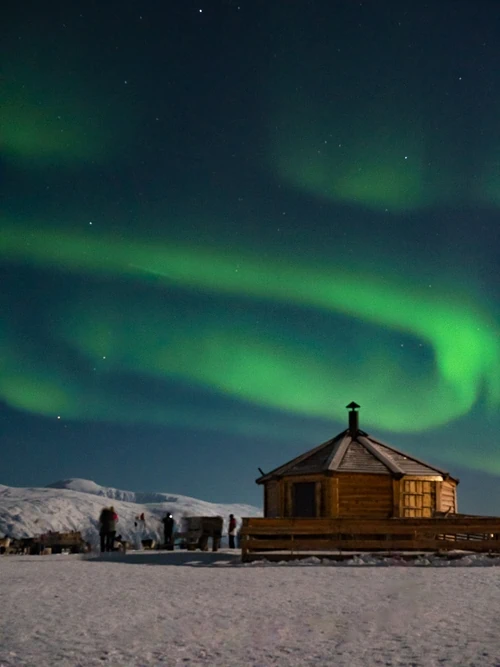 A small wooden hut surrounded by snowy landscapes with a backdrop of night sky full of green Northern Lights, a highlight of this Tromso itinerary
