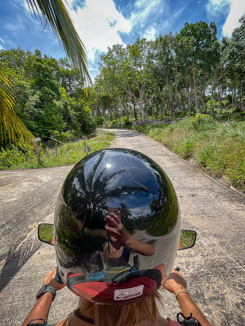 A man wearing a helmet, driving a scooter along a small paved road surrounded by lush forest.
