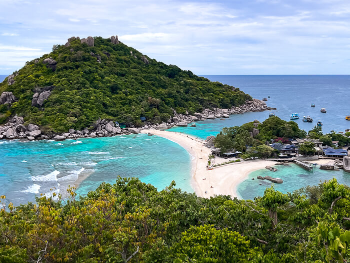 A view over the narrow white sandbank of Koh Nang Yuan island surrounded by tropical blue water and hills covered with forest.