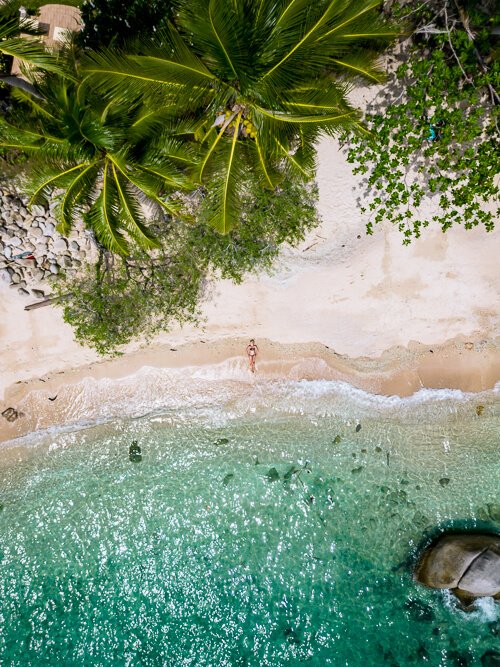 A top-down drone photo of June Juea Beach with its strip of white sand, palm trees and clear turquoise water on Koh Tao island.