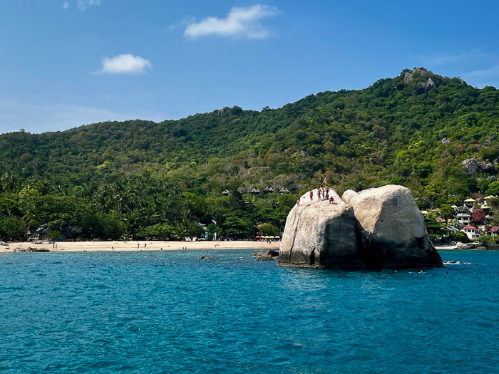 A large rock jutting out of the sea in front of Tanote Bay Beach with a backdrop of forest-covered mountains.