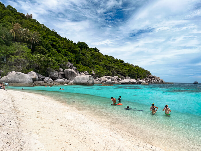 Tourists swimming and snorkeling at a white sand beach with vibrant turquoise water.