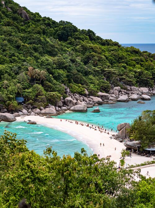 A narrow white sandbank bordered by turquoise water from both sides and surrounded by rugged hills covered with jungle.