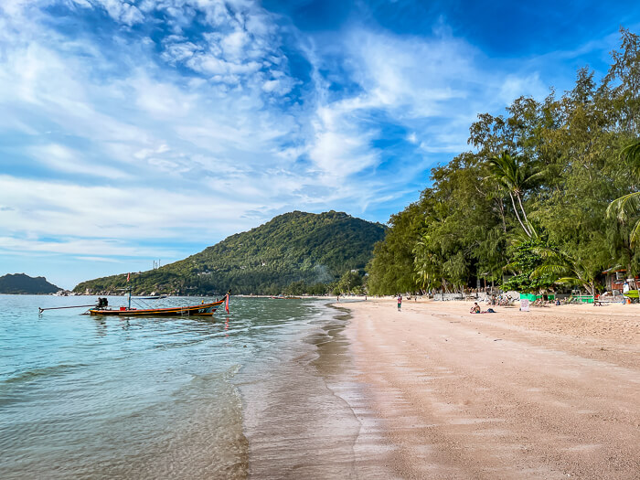 A long strip of tan sand and calm blue sea with a longtail boat on a sunny day at Sairee Beach.