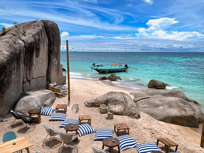 A beach cafe with chairs and small tables on the sand, surrounded by large boulders and overlooking the turquoise sea at Koh Tao
