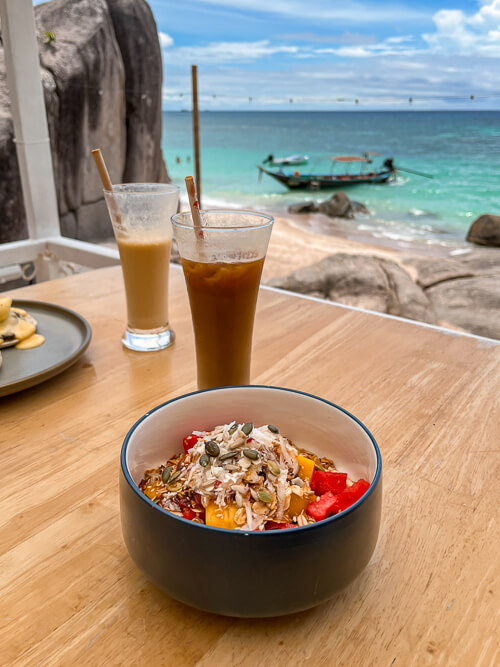 A bowl of granola and fresh fruits with a view of vibrant turquoise sea in the background at Sandbar Beach Club.