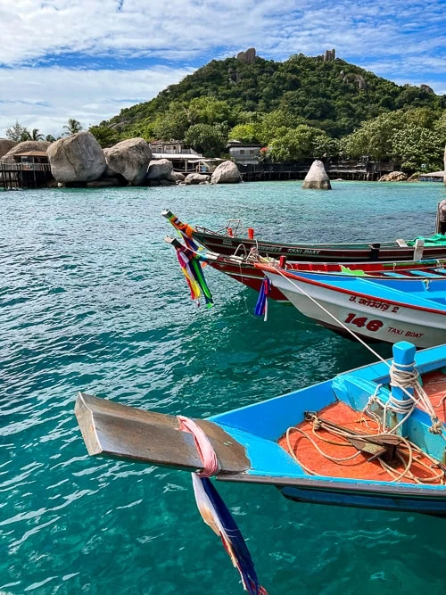 Colorful longtail boats docked next to each other with green forest-covered hills in the background.
