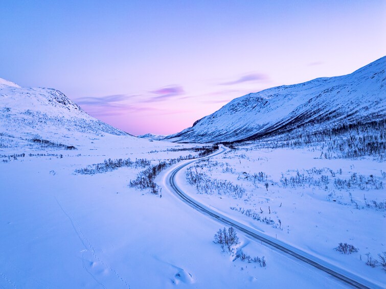 A valley covered with snow and surrounded by mountains with pastel-pink sky in the background on Kvaløya island during the blue hour.