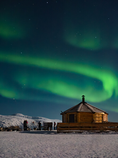 A small wooden hut surrounded by snow with a backdrop of clear night sky full of green Northern Lights near Tromso.