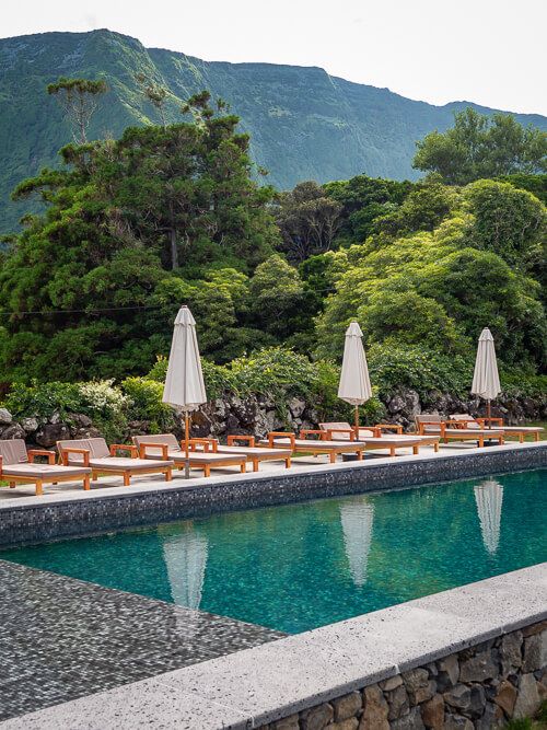 A turquoise swimming pool surrounded by sunbeds and tall green mountains in the background at Aldeia da Cuada hotel.