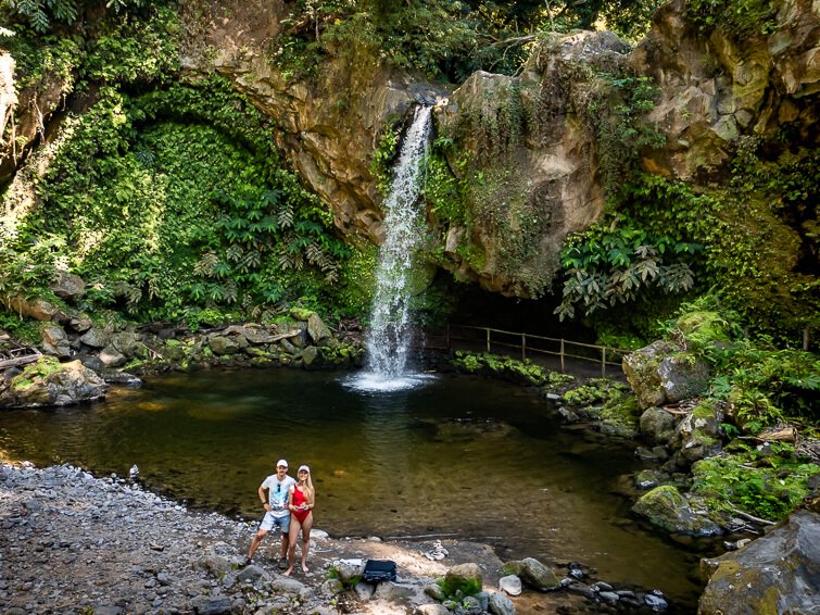 Me and my husband standing in front of the plunge pool of Cascata da Gruta waterfall surrounded by lush plants
