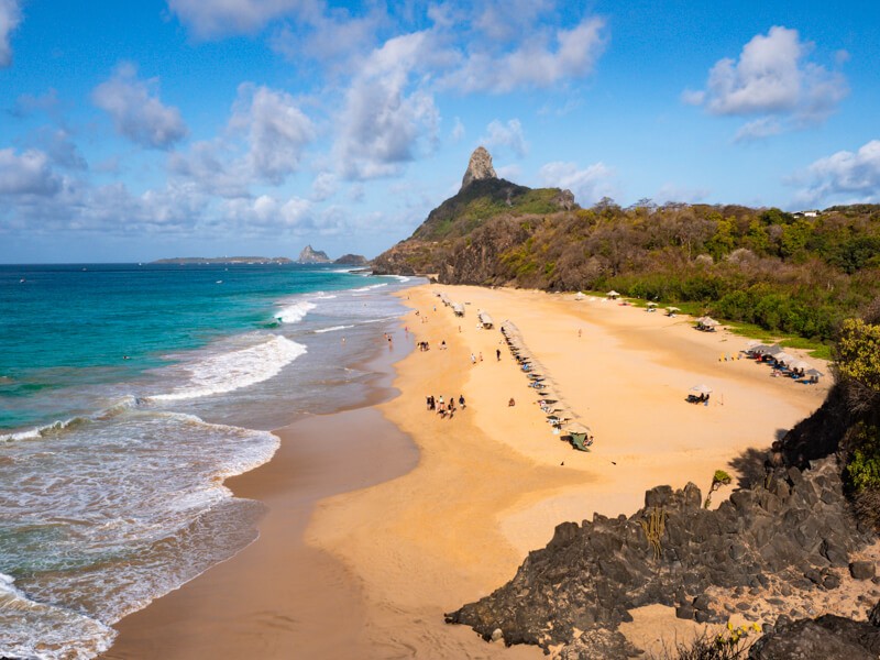 A bird's eye view of Cacimba do Padre Beach, surrounded by green vegetation and volcanic terrain.
