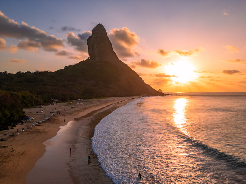 An aerial photo of Conceicao Beach with its long strip of sand and the tall pointy Pico mountain in the background during sunset.