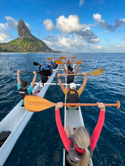 Our group posing with their hands and oars in the air during our Hawaiian canoe tour, with the Morro do Pico mountain in the background.