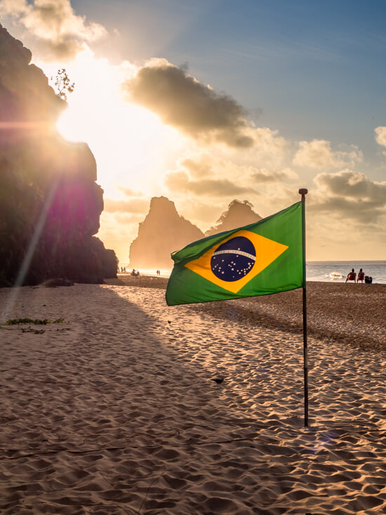A Brazilian flag waving in the wind on a large sandy beach with the rays of sun in the background.