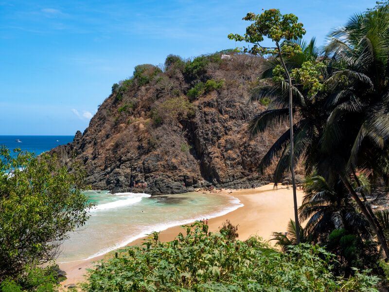 The small sandy Praia do Cachorro Beach surrounded by green vegetation and a large rugged hill in the background.