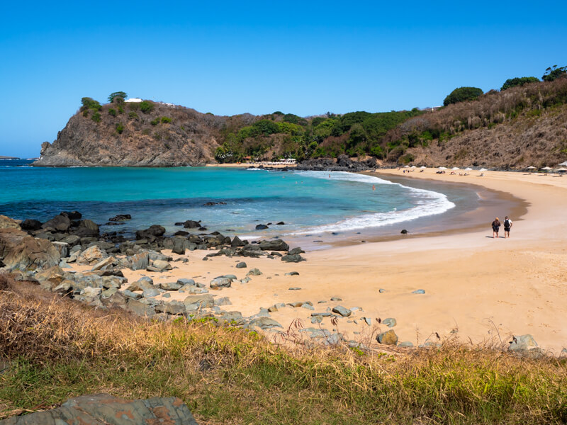 The small quiet sandy beach of Praia do Meio surrounded by hills covered with dry vegetation.
